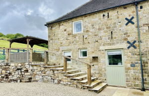 A high-end barn conversion living area featuring vaulted ceilings, exposed timber beams, sleek modern furniture, and large glass doors leading to a private patio.